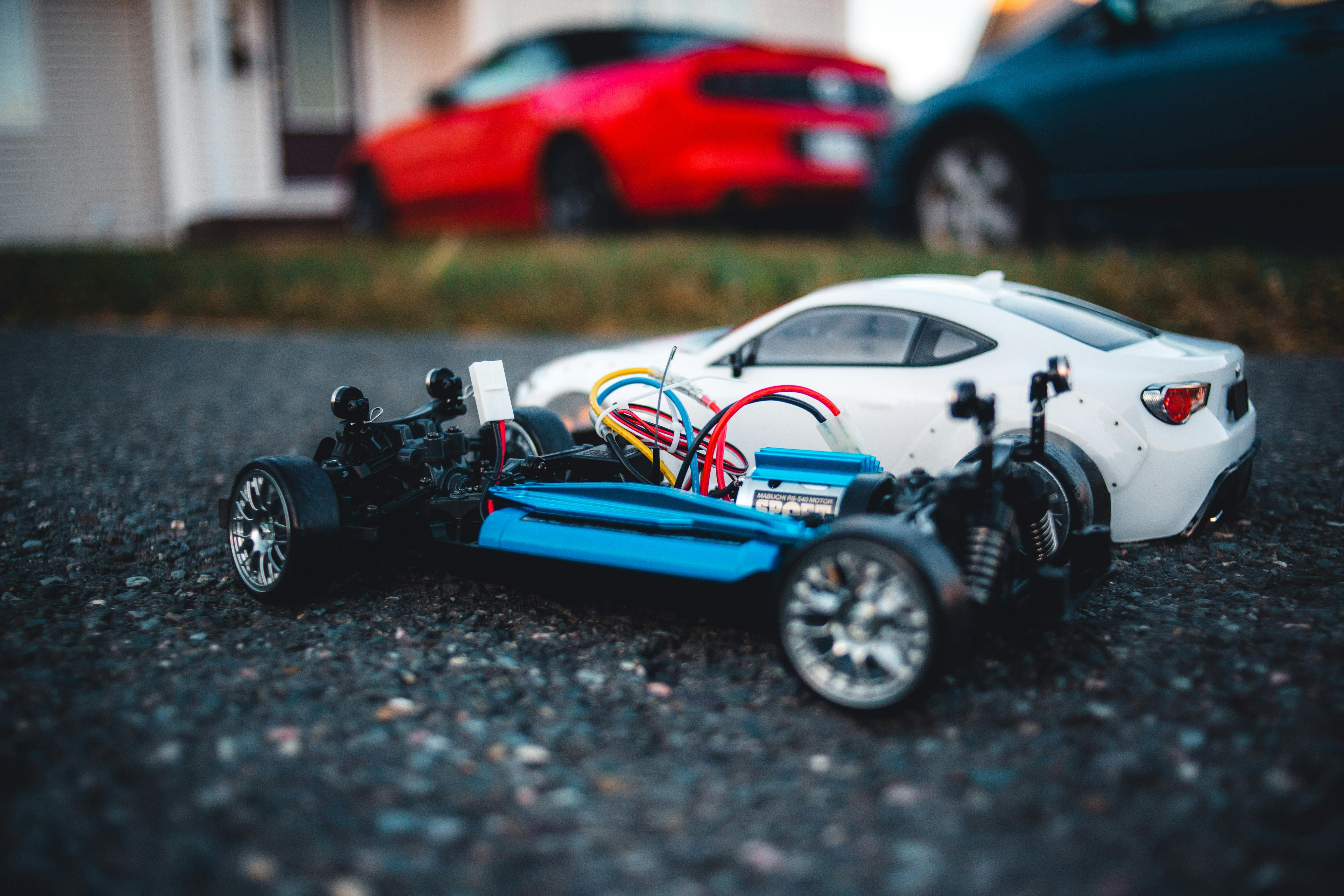 Close-up of two RC cars on asphalt with full-sized cars blurred in the background.
